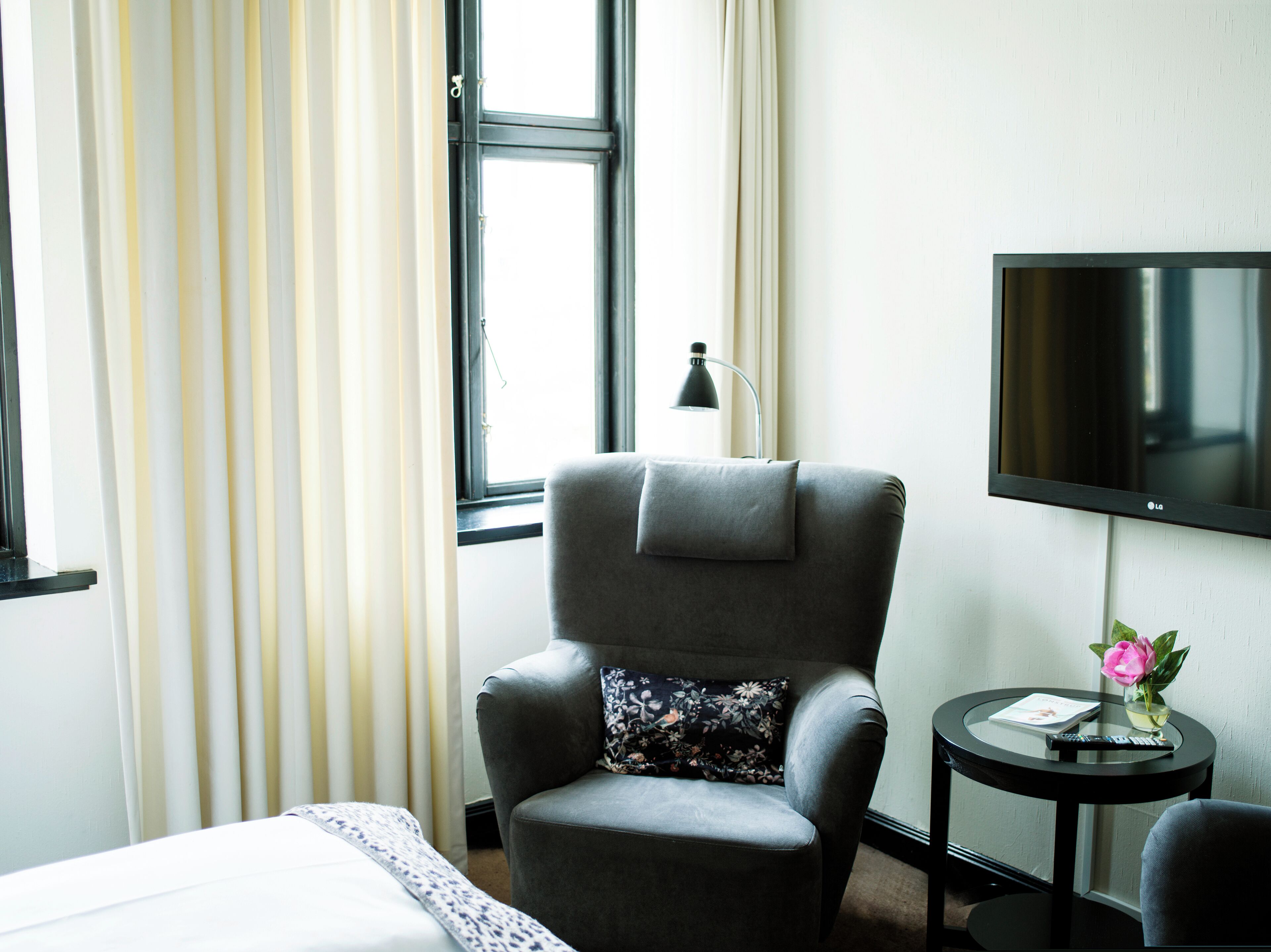 A hotel room with a grey armchair, a small round table with a remote control, a flower vase, and a TV on the wall.