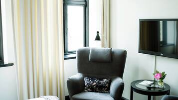 A hotel room with a grey armchair, a small round table with a remote control, a flower vase, and a TV on the wall.