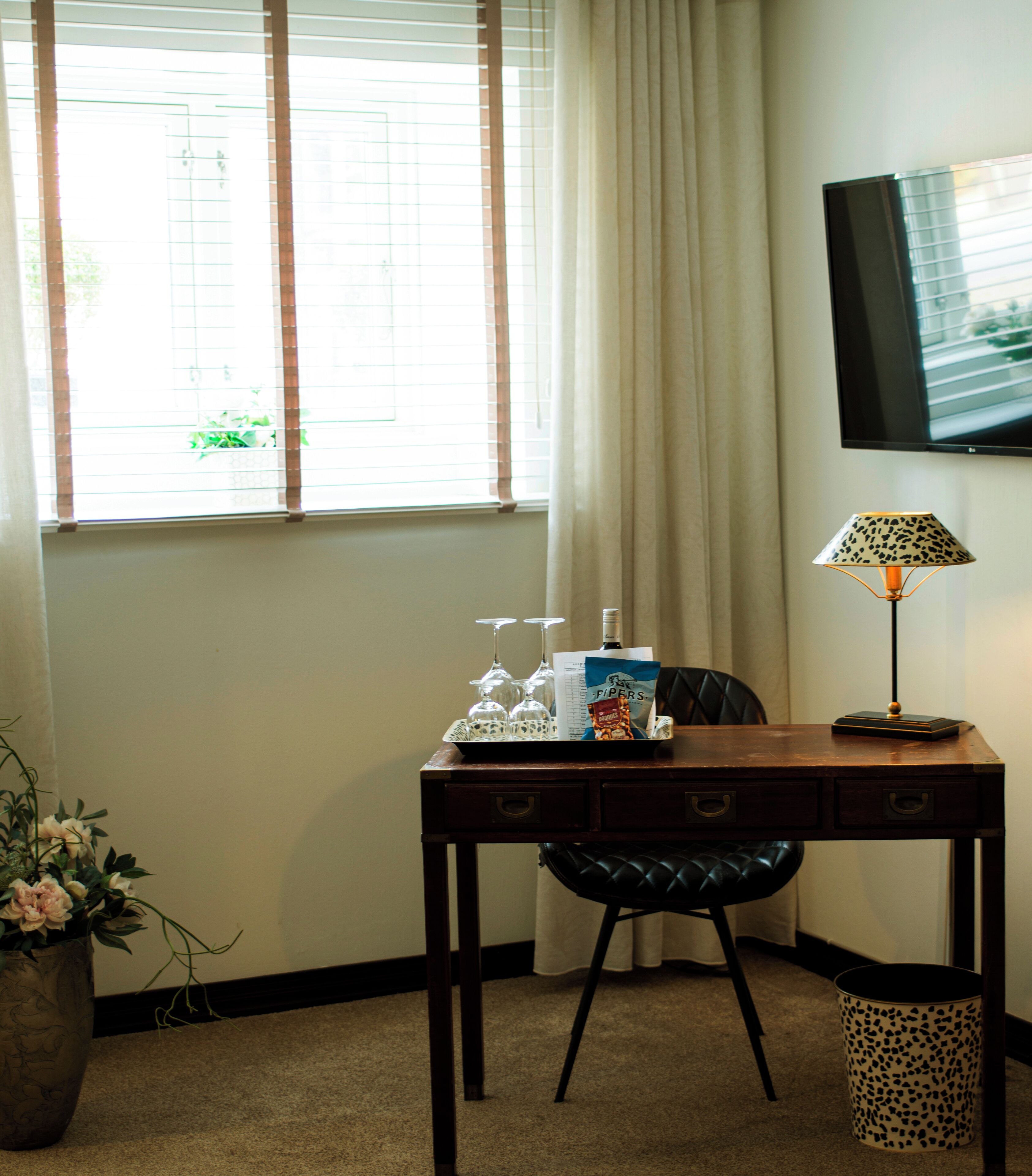 A hotel room with a wooden desk, a chair, a television mounted on the wall, a window with blinds, and a potted plant.