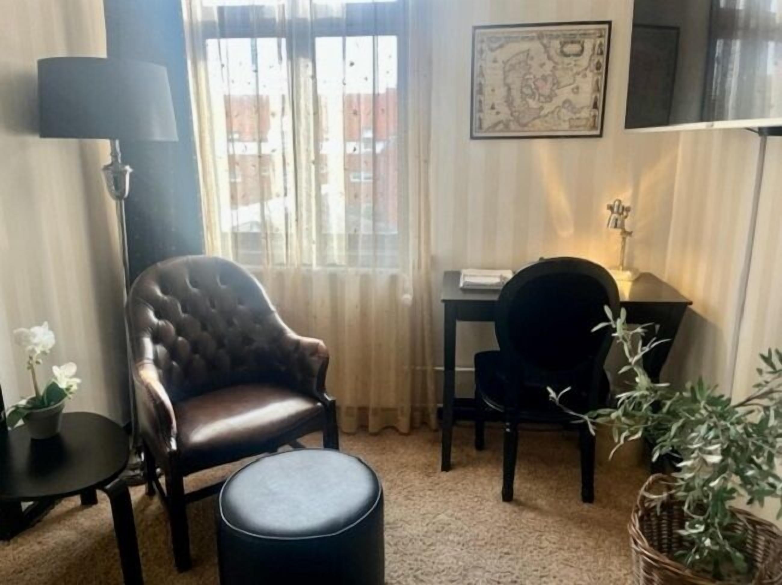 A room with a leather armchair, a small round ottoman, a desk with a chair, a lamp, and a framed map on the wall.