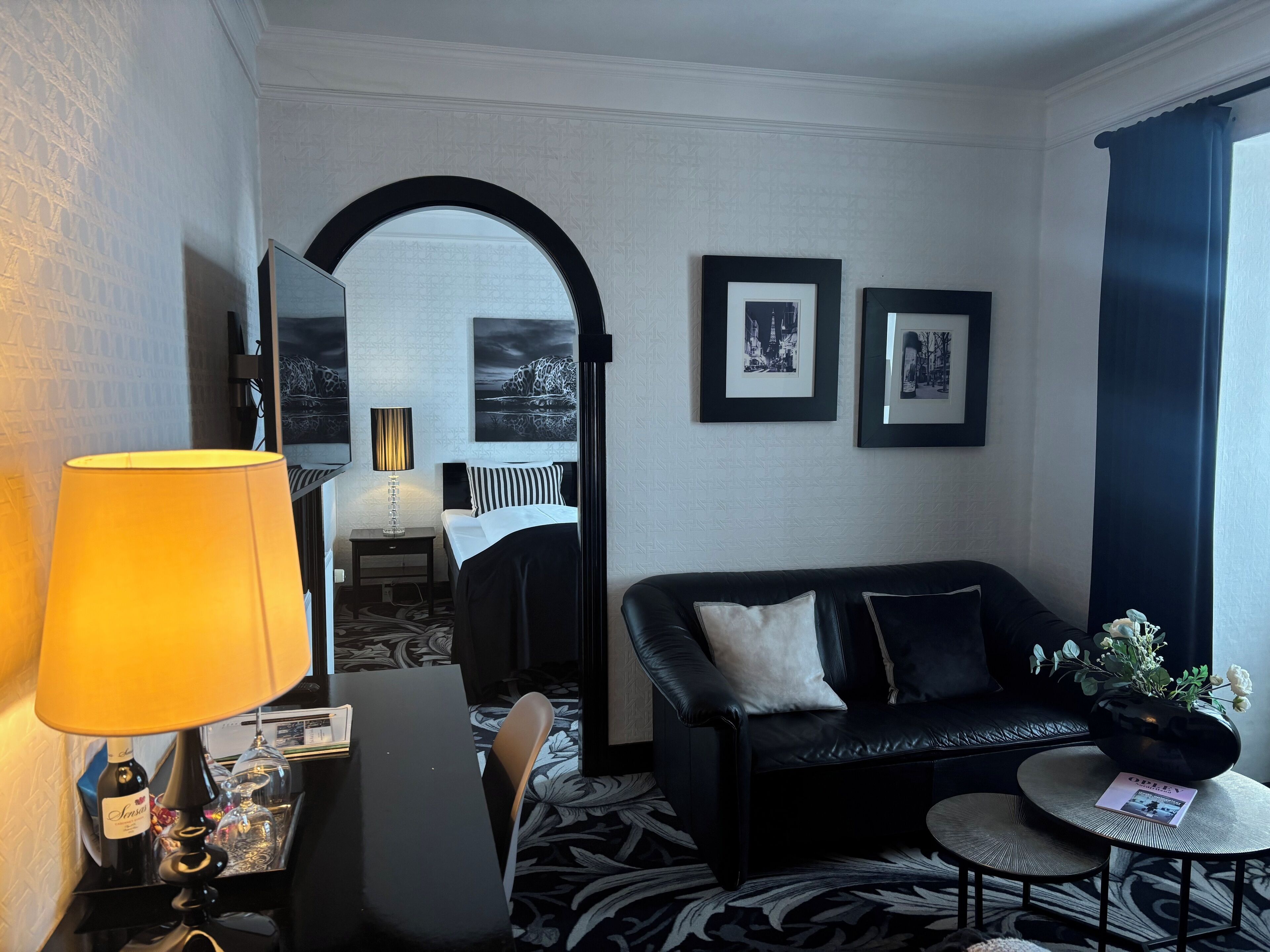A hotel room with a bed, a black leather sofa, a small round table, a lamp, and framed pictures on the wall.