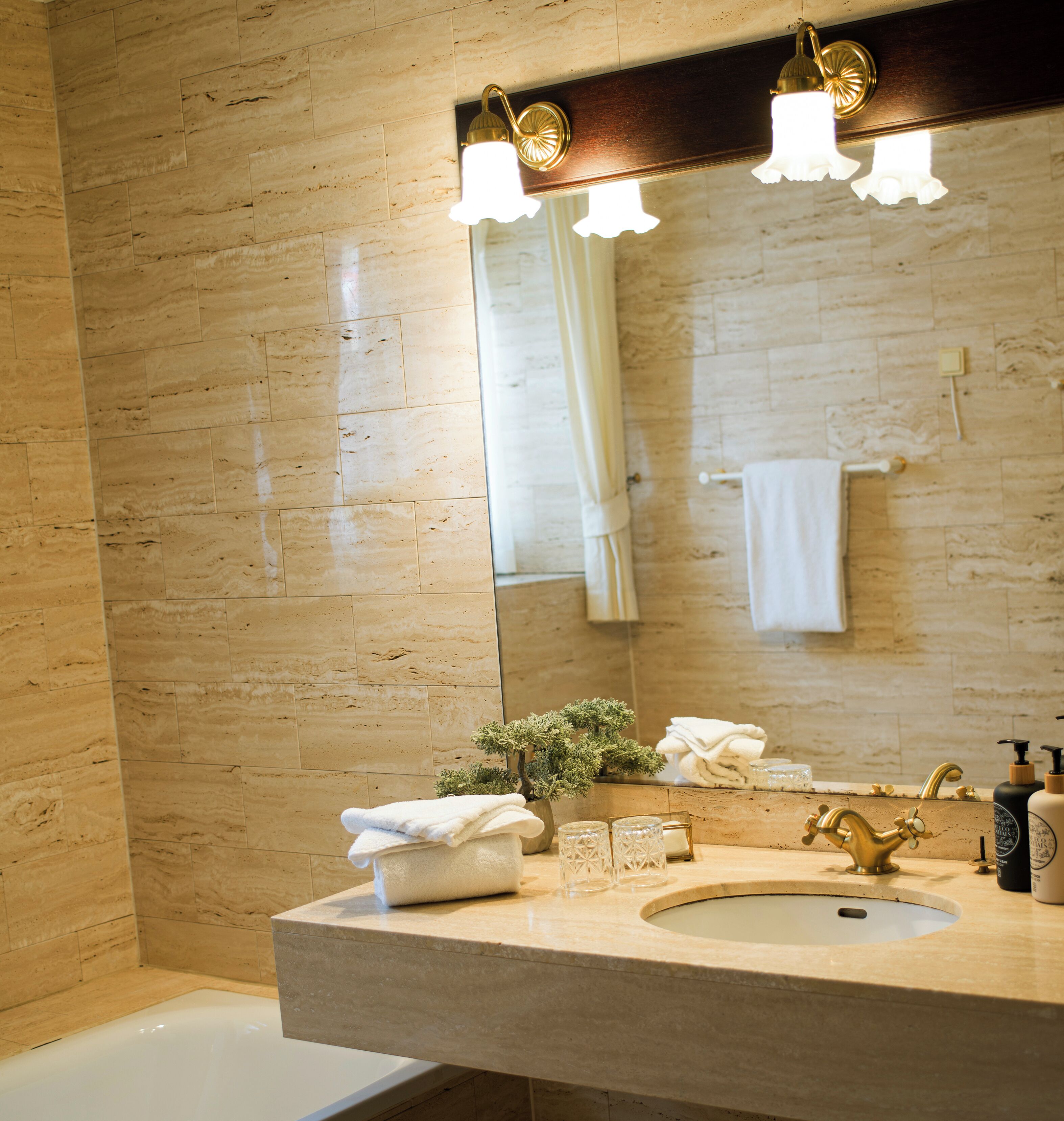 Un baño con pared de mármol, un lavabo con grifo de oro y un espejo sobre el lavabo.