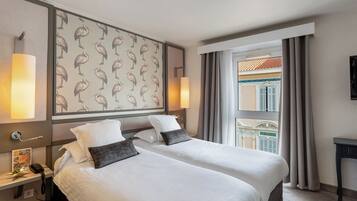 Quarto de hotel com duas camas, mesa de cabeceira com telefone, parede com padrão de pássaros e vista para um edifício pela janela.