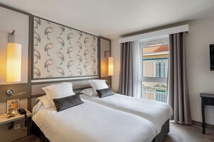 Een hotelkamer met twee bedden, een nachtkastje met een telefoon, een wand met een vogelmotief en uitzicht op een gebouw door een raam.