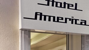 Entrada de un hotel con un cartel que dice "Hotel America", un reloj de pared grande y una zona de estar con una lámpara y sillas.