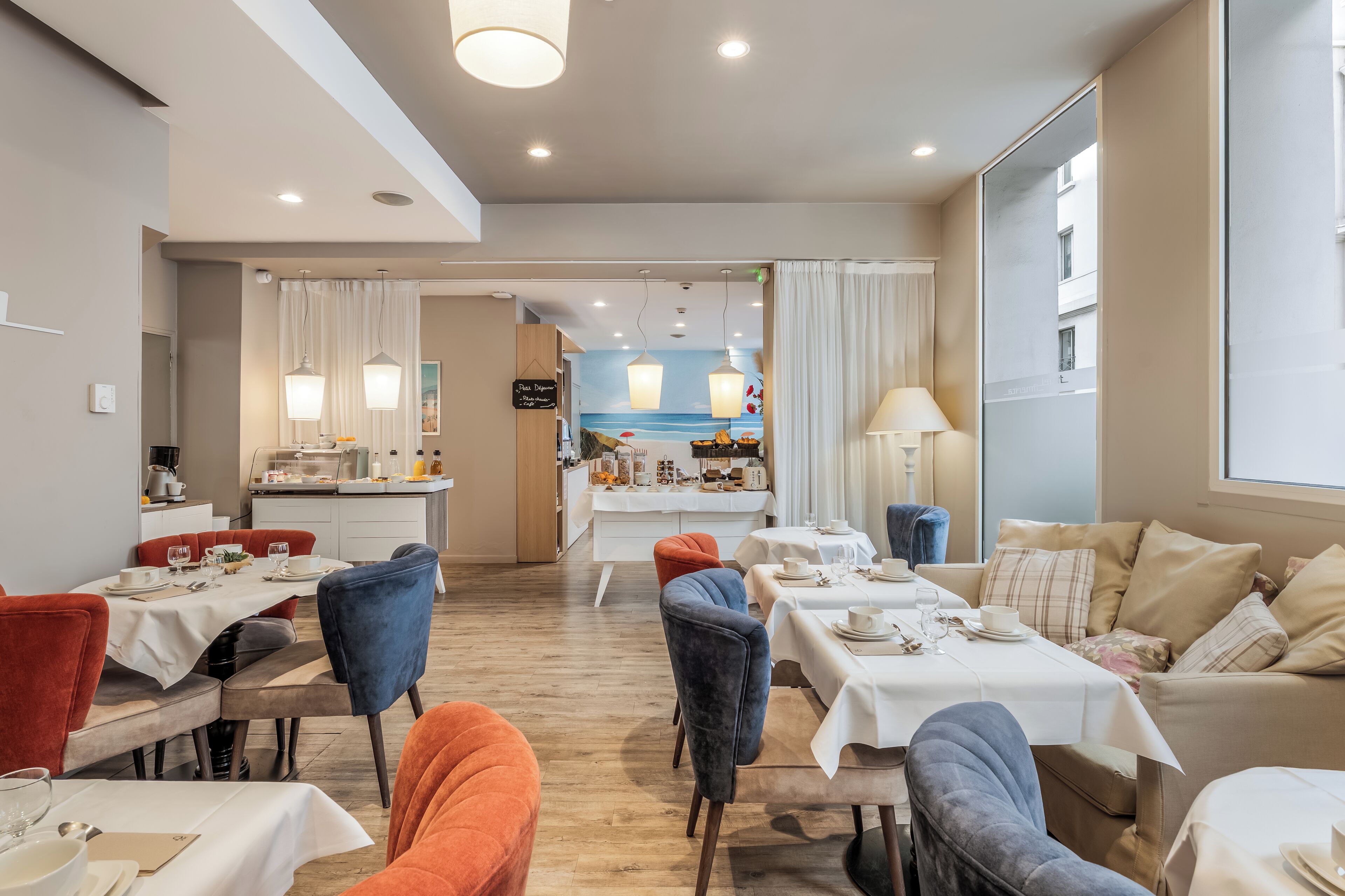 Un comedor de hotel con varias mesas y sillas, una cocina al fondo y vista al mar a través de un amplio ventanal.