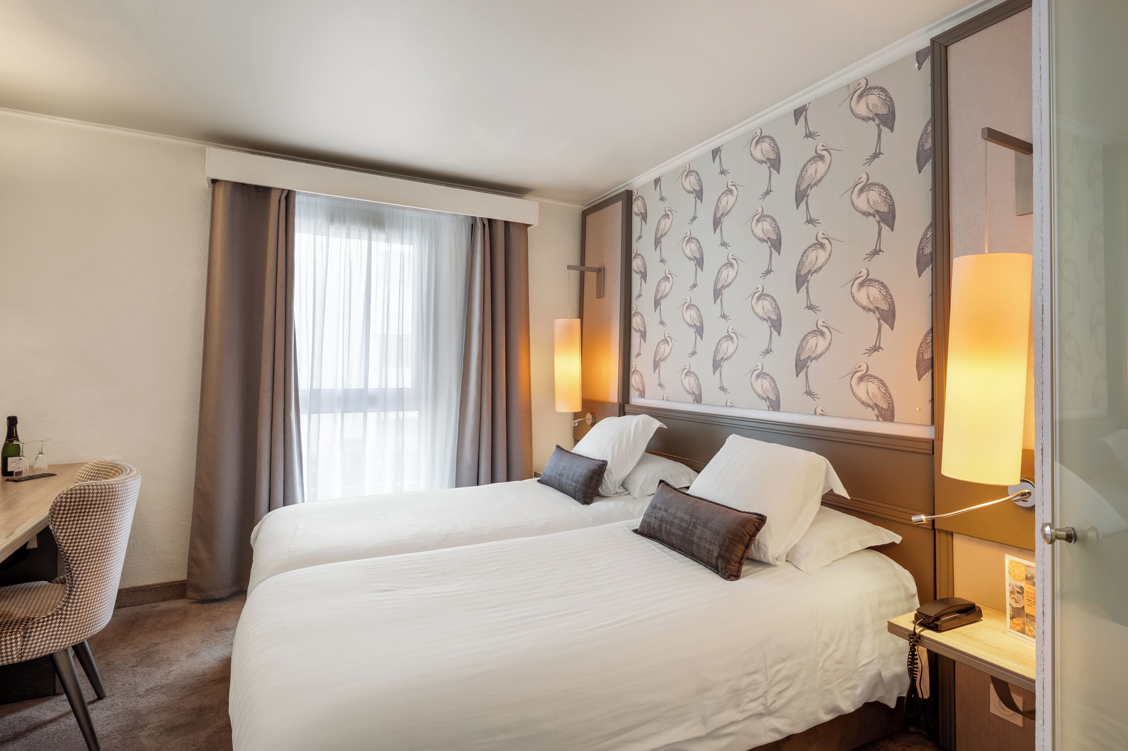 Habitación de hotel con dos camas, un escritorio con silla, un minibar y un papel tapiz con diseño de pájaros.