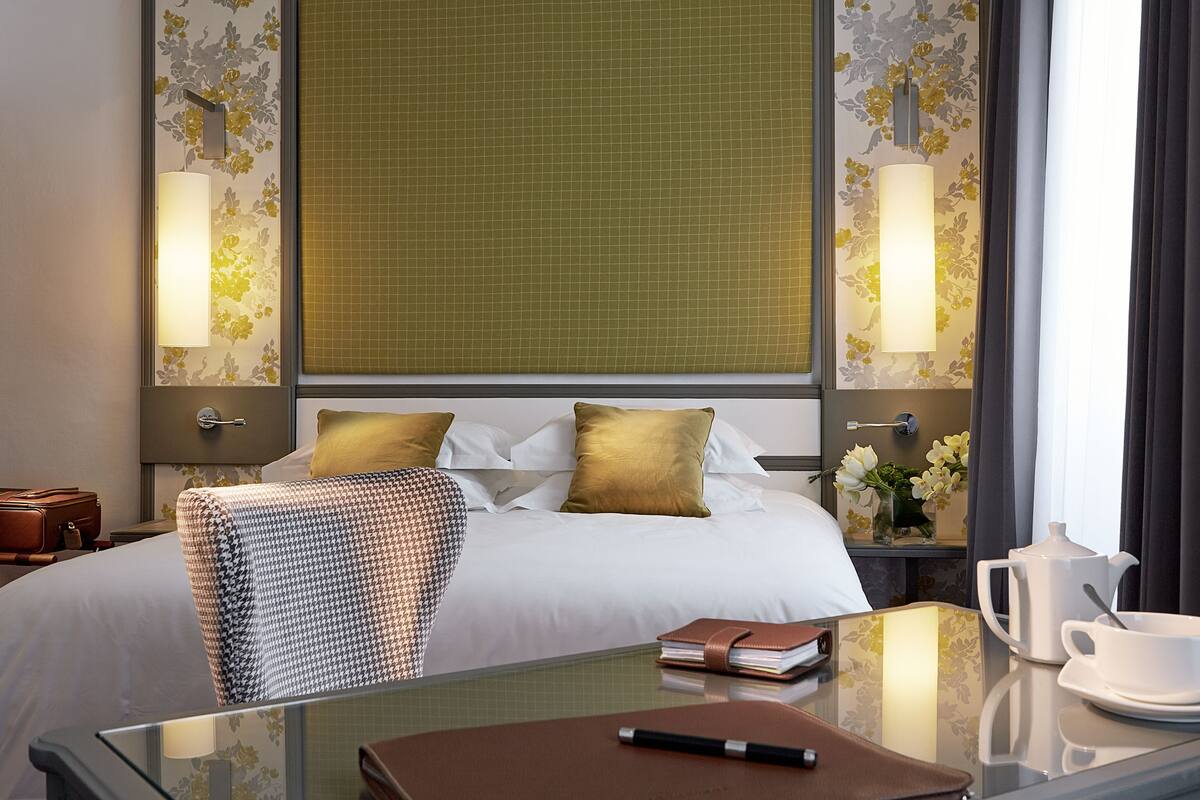 Une chambre d’hôtel avec un grand lit, une chaise, une table avec un carnet, un stylo, une théière et une tasse.