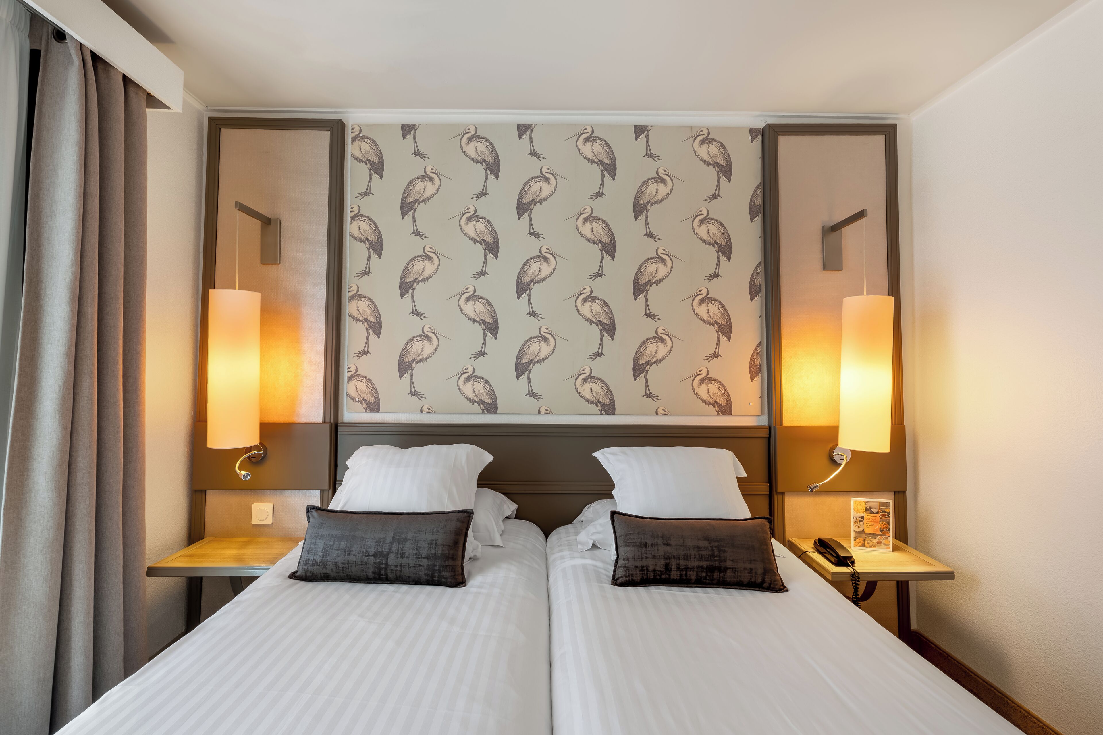 A hotel room with two beds, a headboard with a patterned wallpaper, bedside lamps, and a nightstand with a phone.