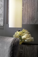 Een lamp met een witte kap, een boeket witte bloemen en een kussen met een hondentandpatroon op een houten ondergrond.