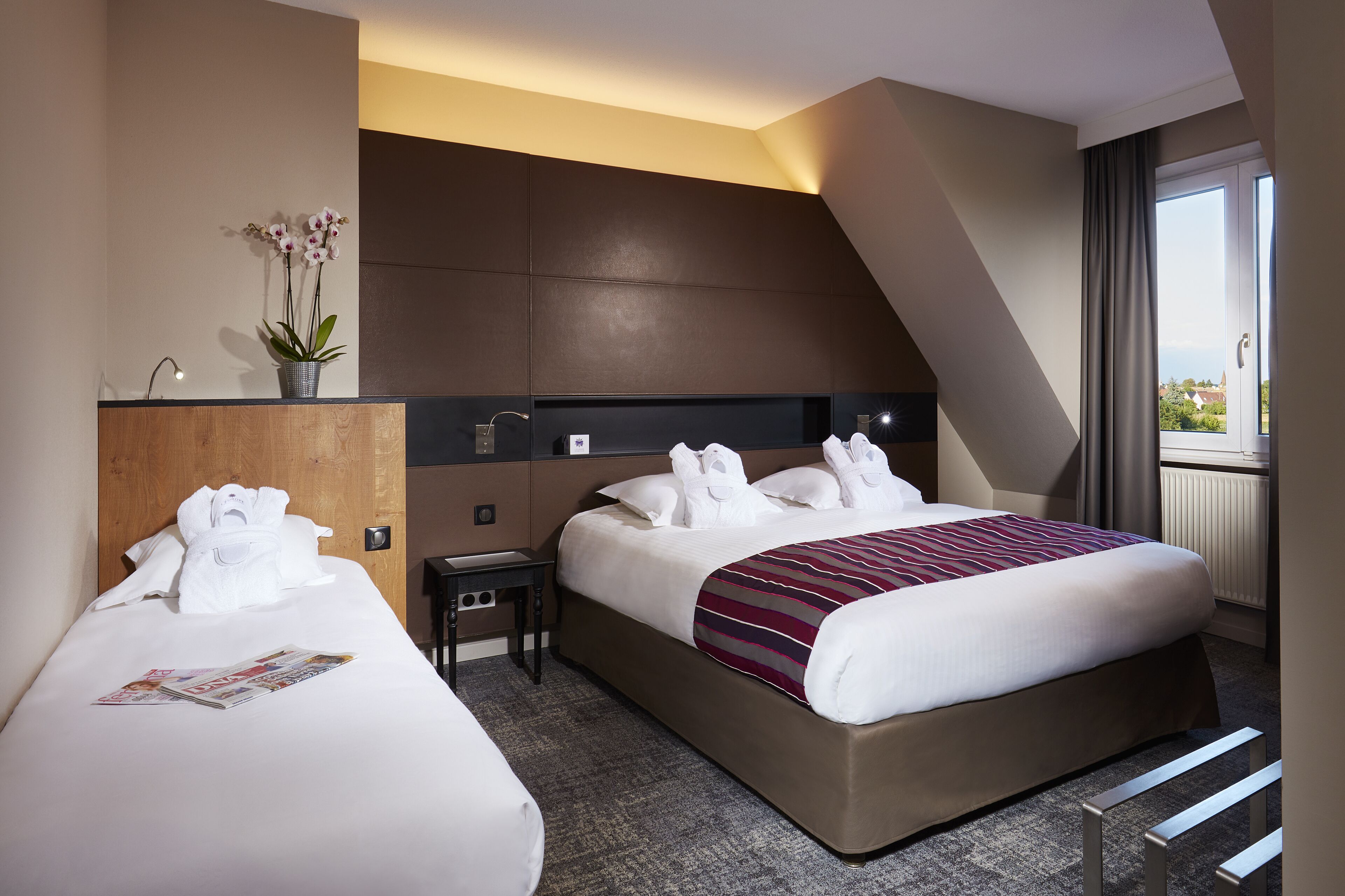 A hotel room with two beds, a bedside table, a lamp, a vase with flowers, and a window with curtains.