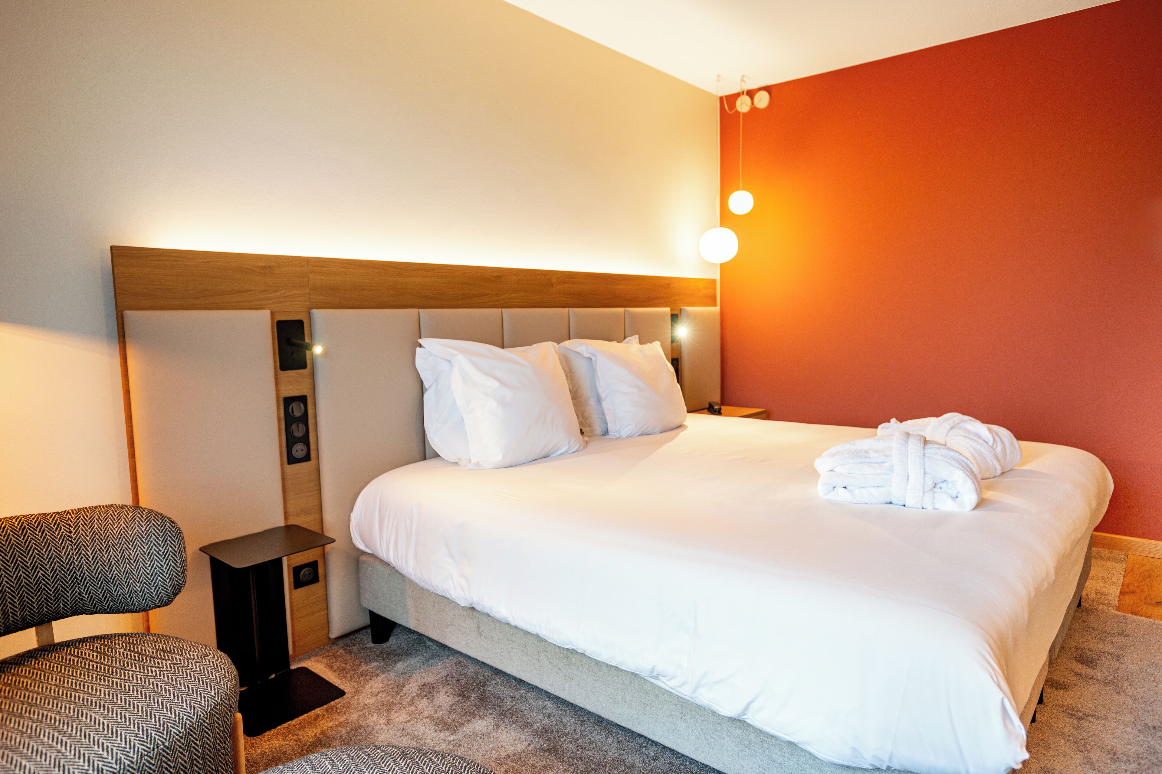 A hotel room with a large bed, a nightstand, a chair, and a wall-mounted light fixture.