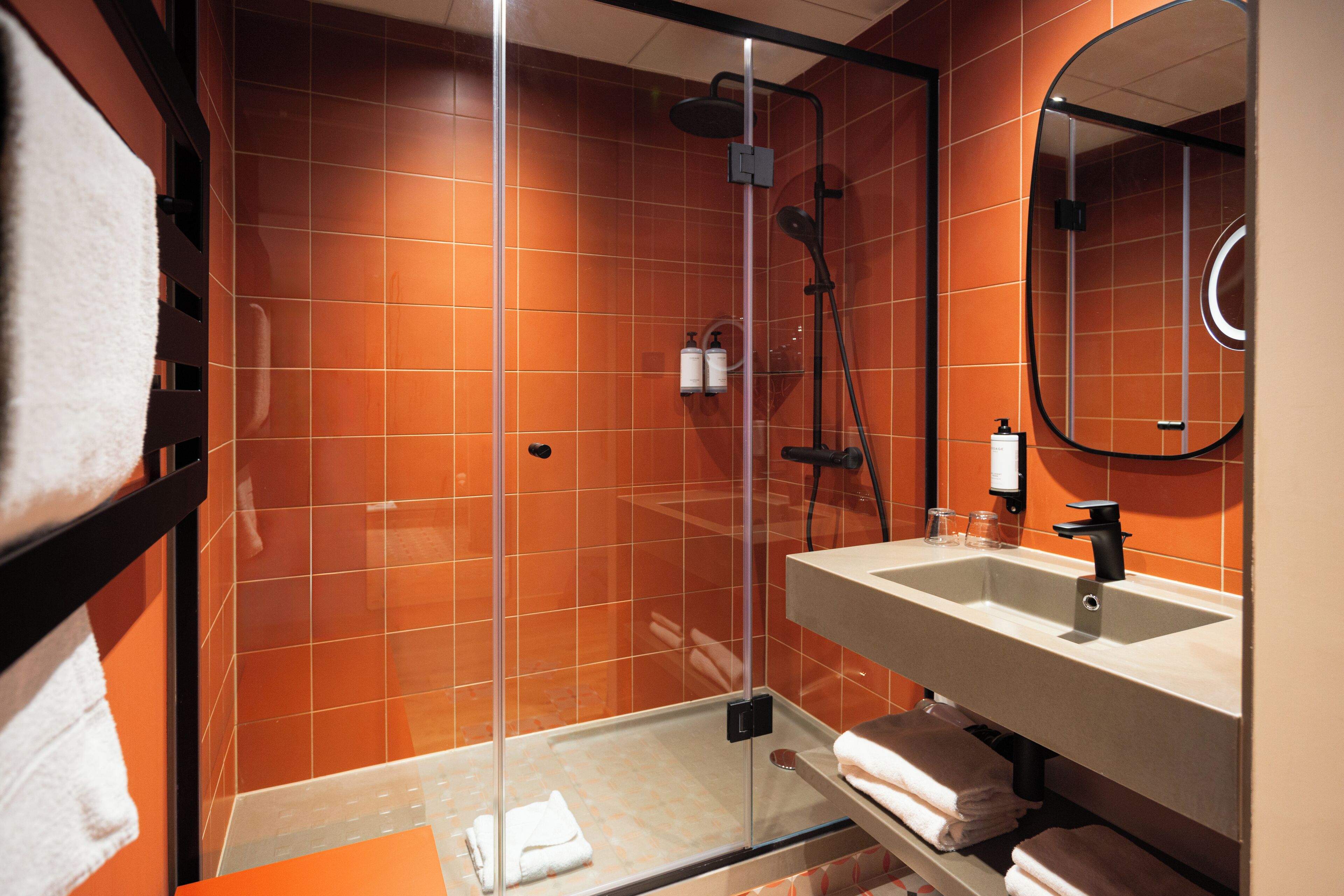 A modern bathroom with orange tiles, a glass-enclosed shower, a sink with a mirror, and neatly folded towels.