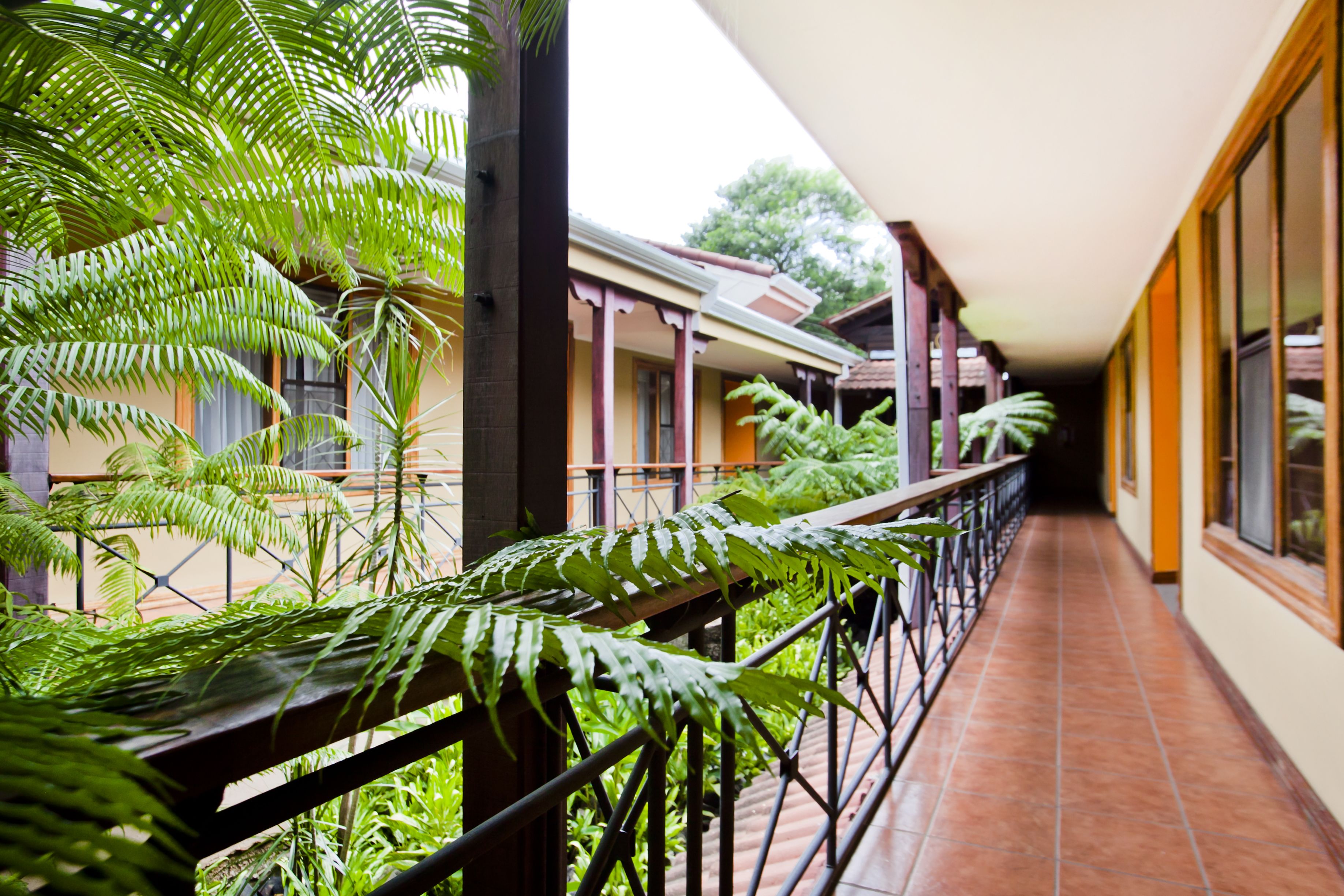 A corridor with a tiled floor, a railing with greenery, and a building with a tiled roof and windows.