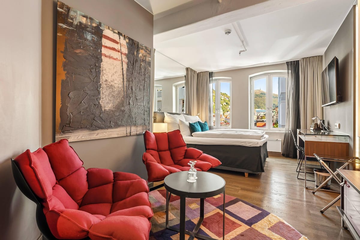 Quarto de hotel moderno com cama, duas cadeiras vermelhas, uma mesinha, um quadro abstrato grande, televisão e varanda com vista.