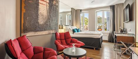 A modern hotel room with a large abstract painting, a bed with a view, a desk, and two red armchairs.