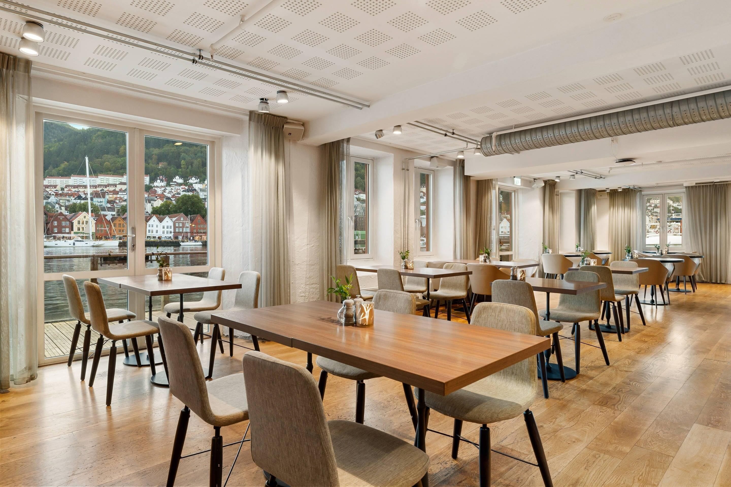 A spacious dining area with multiple tables and chairs, large windows offering a view of a town and water, and a high ceiling with recessed lighting.