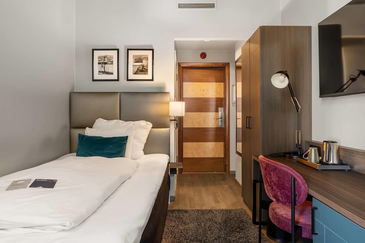 Quarto de hotel com cama, escrivaninha, cadeira, abajur e porta.