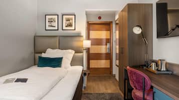 A hotel room with a bed, a desk, a chair, a lamp, and a door.