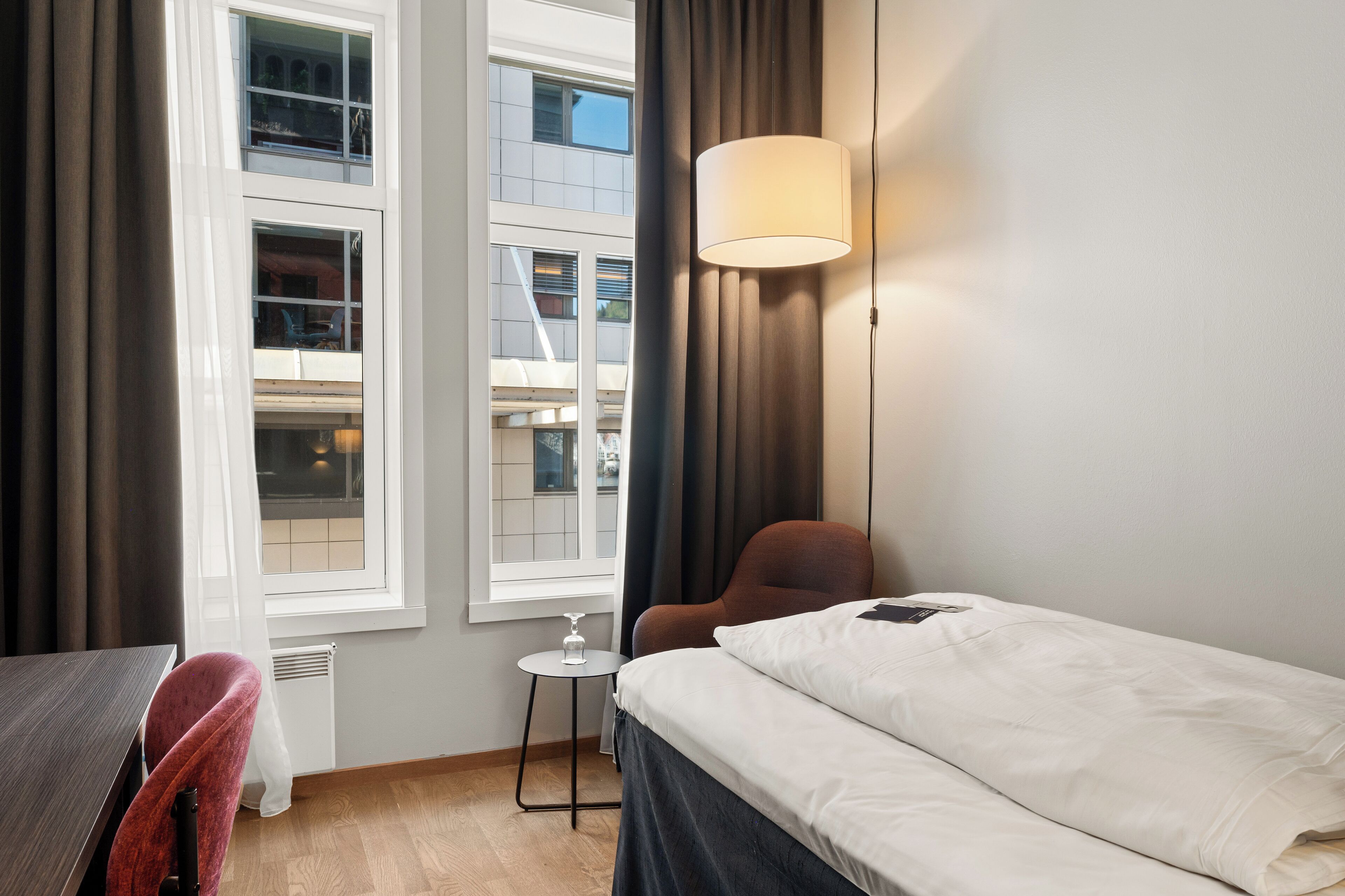 Quarto de hotel com cama, cadeira, mesinha e janela com cortinas.