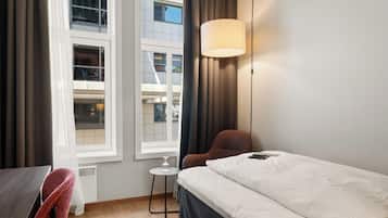 Quarto de hotel com cama, cadeira, mesinha e janela com cortinas.