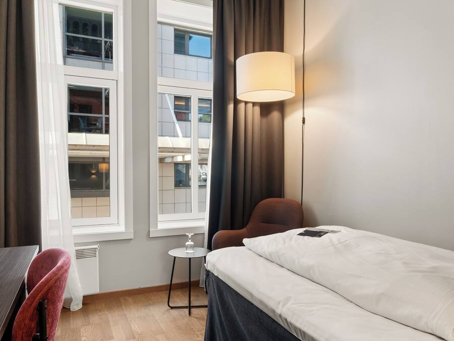 Quarto de hotel com cama, cadeira, mesinha e janela com cortinas.
