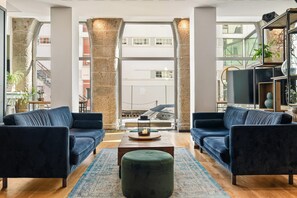 A modern living room with two dark sofas, a green ottoman, and a coffee table. Large windows provide a view of the cityscape.
