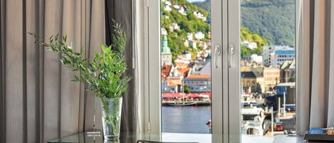 A room with a desk, a vase with green plants, and a view of a town by the water.