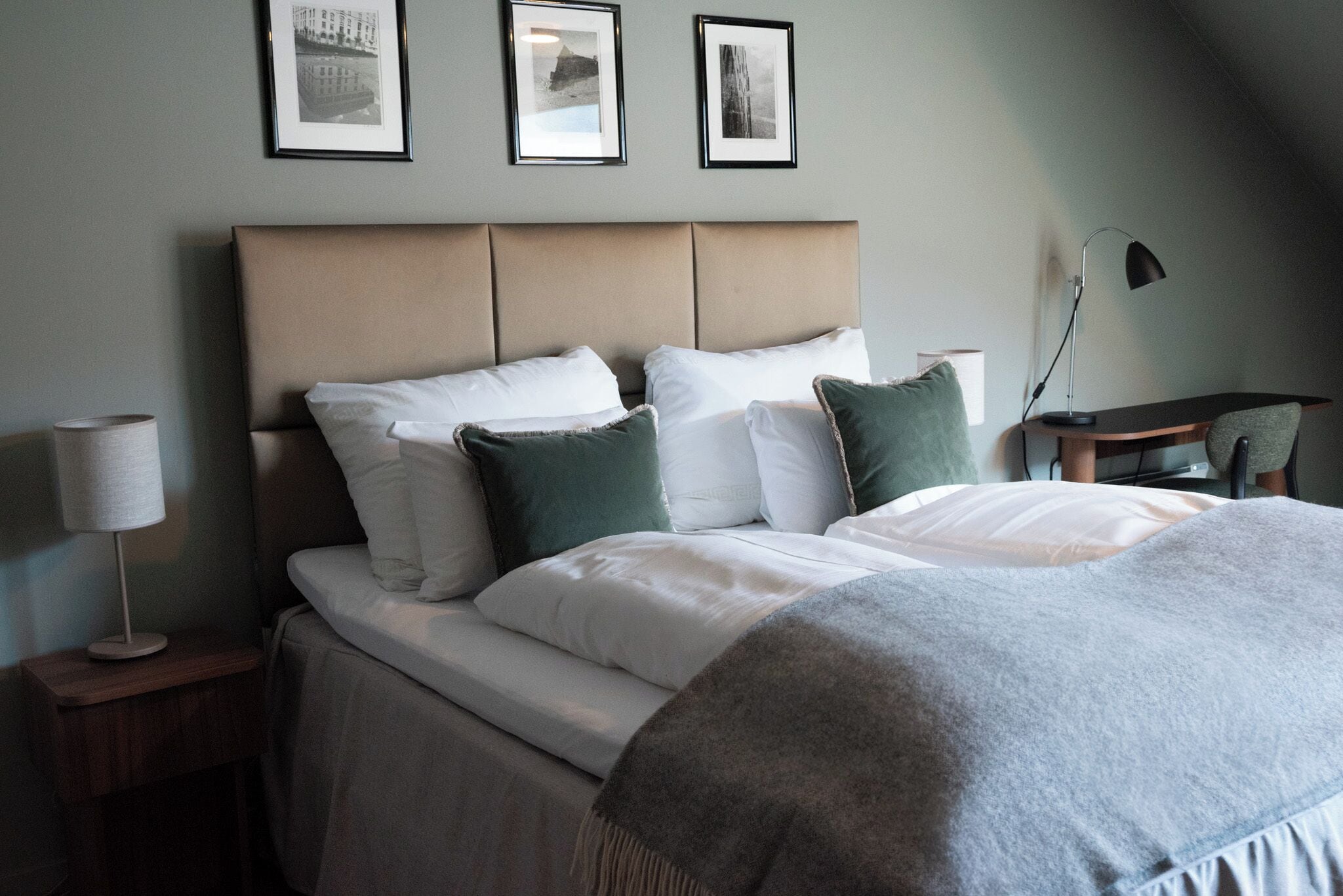A neatly made bed with white and green pillows, a grey throw blanket, and a headboard with three framed pictures on the wall.