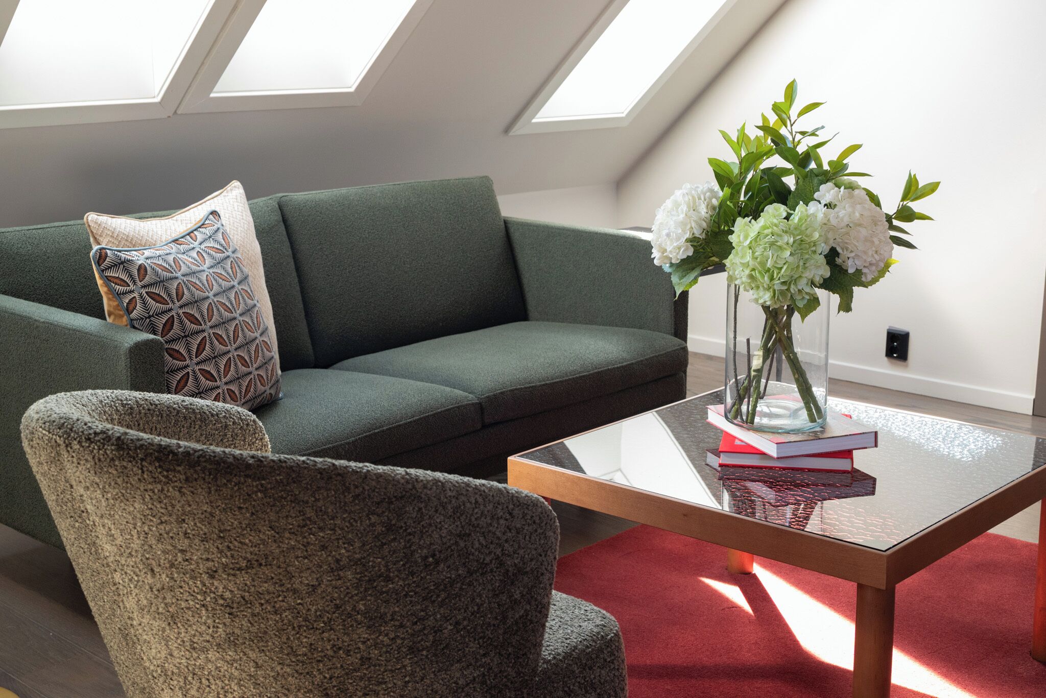 A modern living room with a green sofa, a patterned pillow, a glass coffee table with books and a vase of flowers, and skylights.