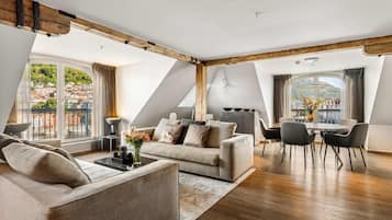 A modern loft apartment with a high ceiling, exposed wooden beams, a sofa, a dining table with chairs, and large windows offering views of a town and mountains.