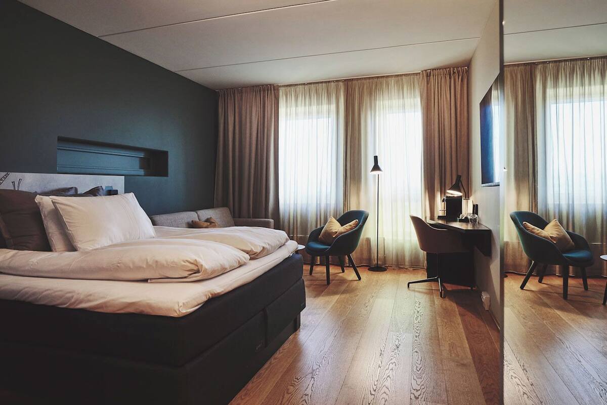 Une chambre d’hôtel moderne avec un grand lit, deux fauteuils, un bureau et une grande fenêtre avec des rideaux.