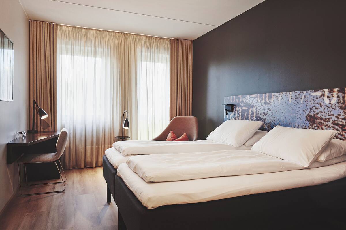 Une chambre d’hôtel avec deux lits, un bureau, une chaise et une grande fenêtre avec des rideaux.