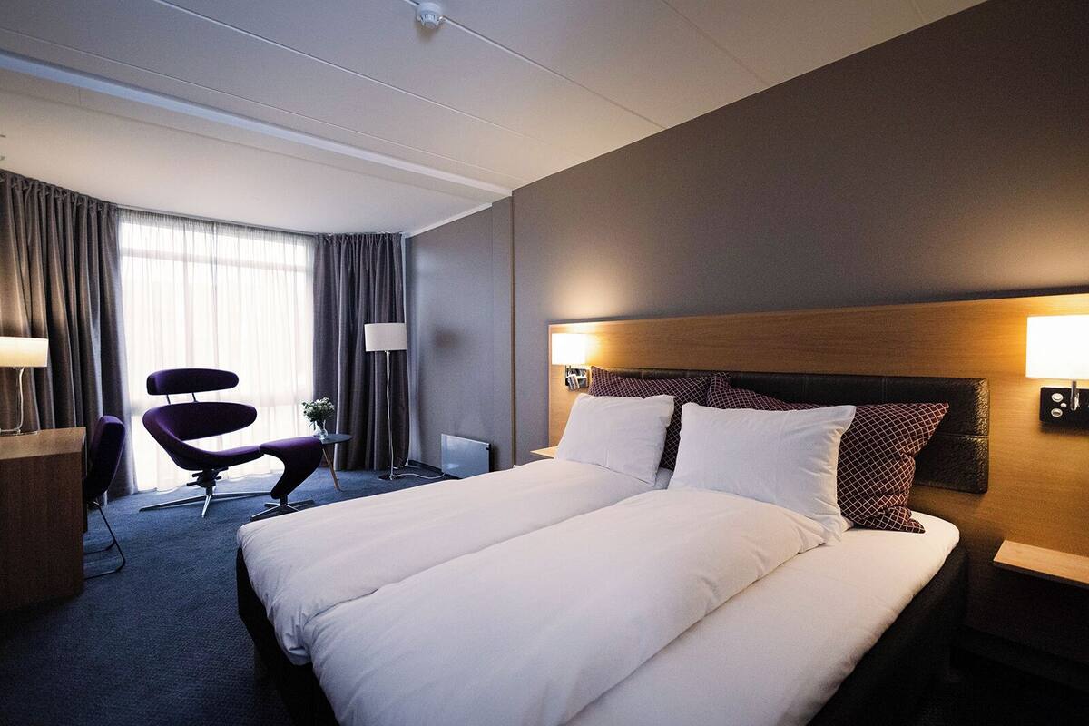 Une chambre d’hôtel avec un grand lit, un bureau, une chaise, une lampe et une fenêtre avec des rideaux.