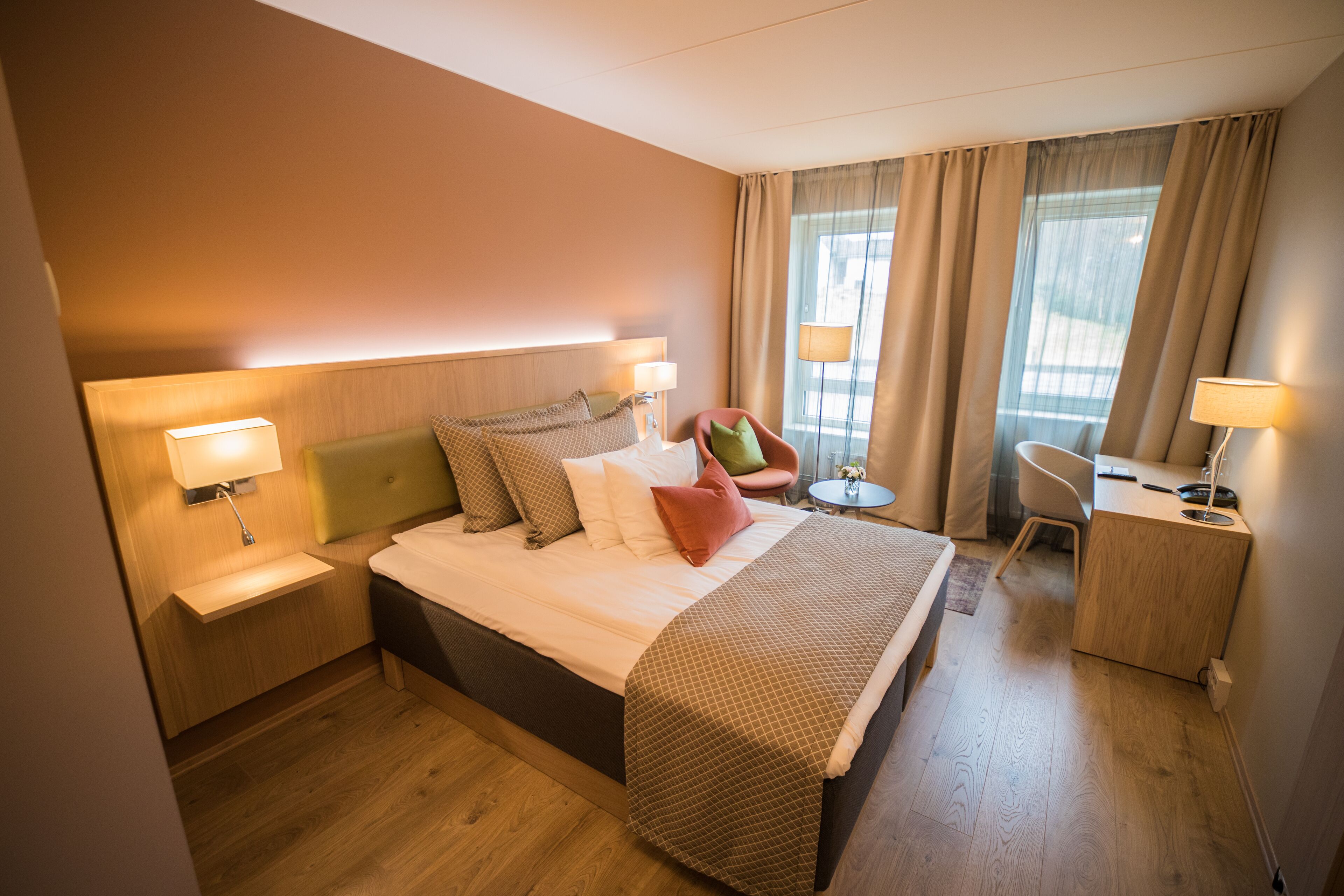 A hotel room with a bed, bedside tables, a desk, a chair, and a window with curtains.