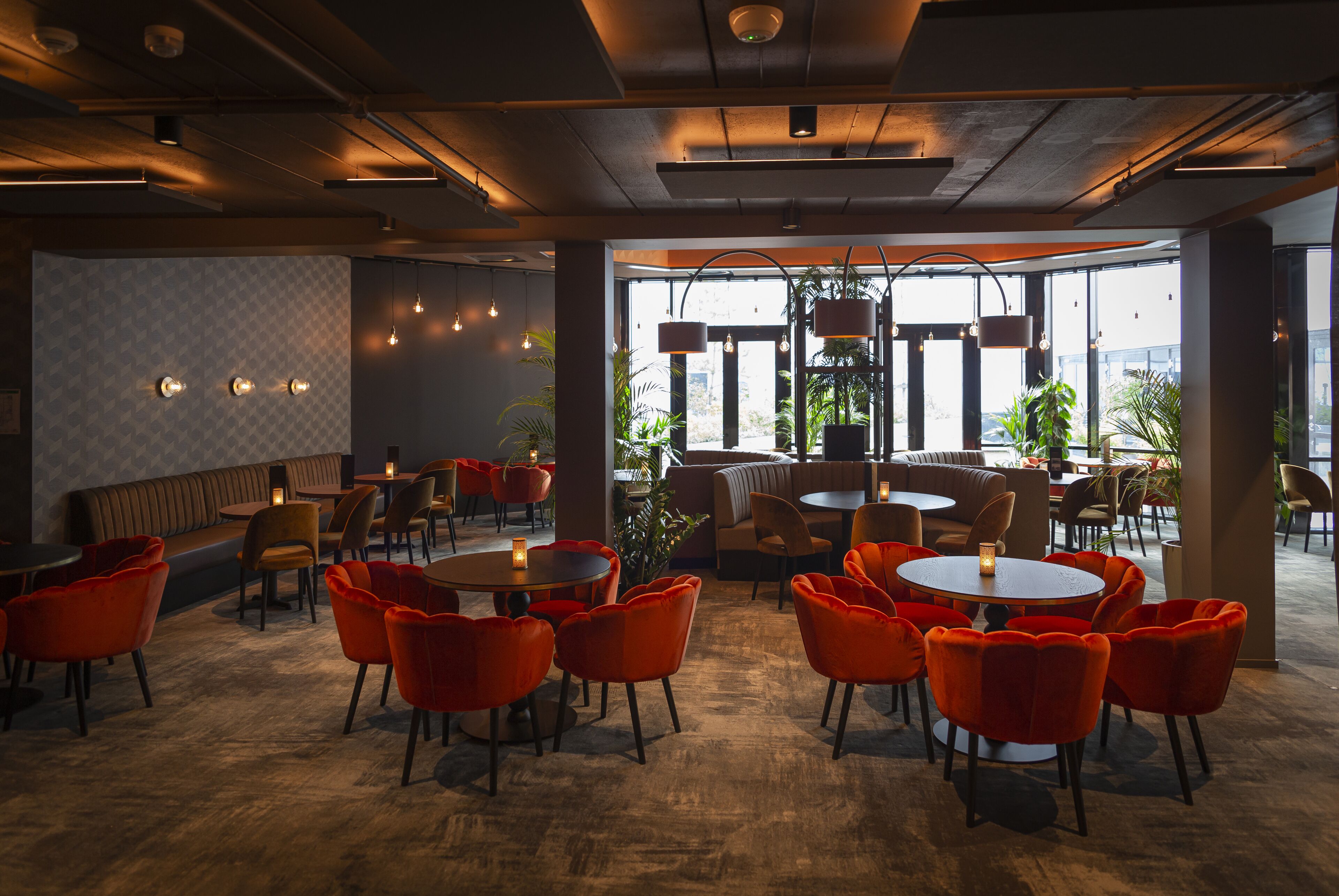 A modern restaurant with round tables and orange chairs, featuring a view of the city through large windows.