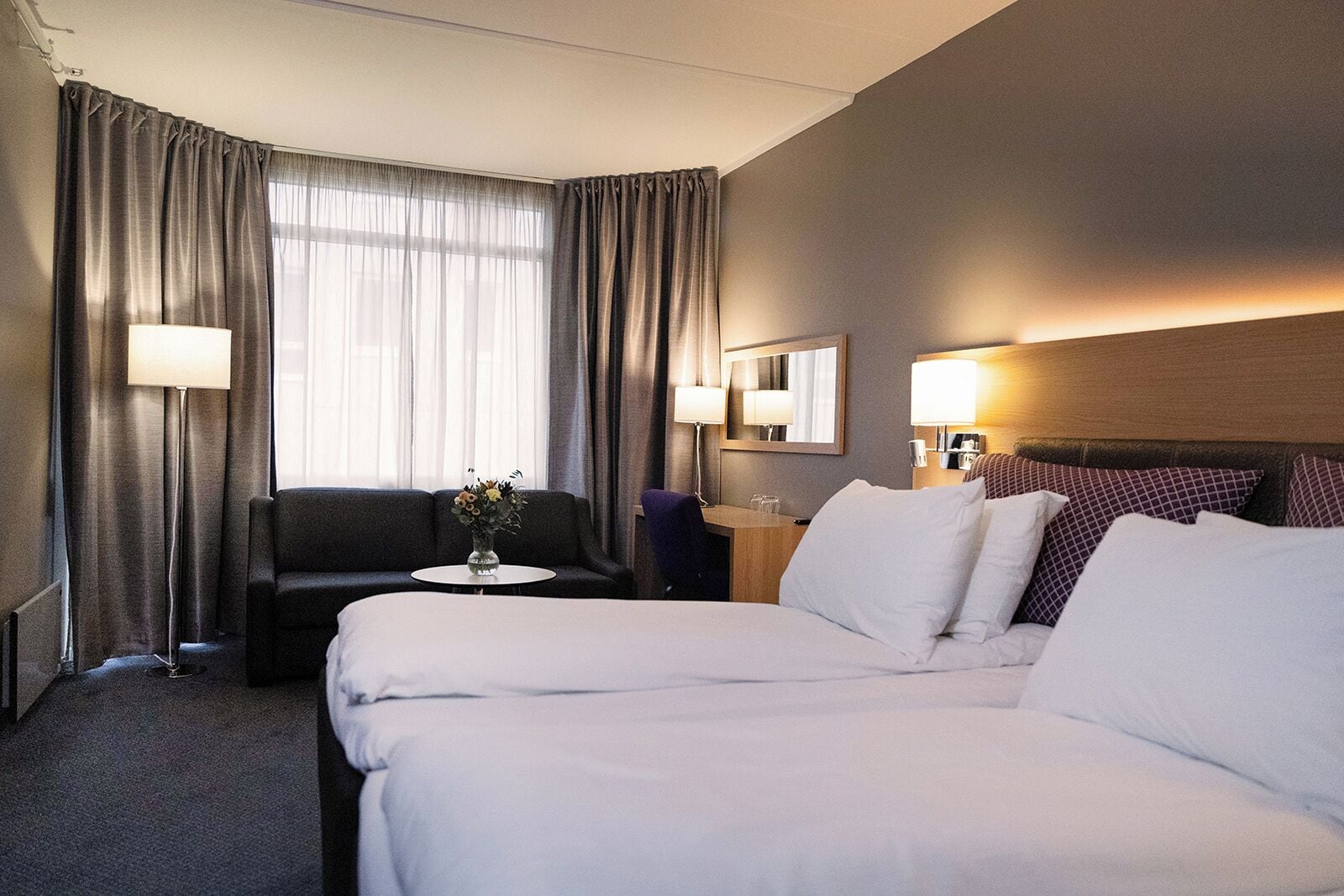 A hotel room with a large bed, a sofa, a small table with a vase, and a desk with a lamp.