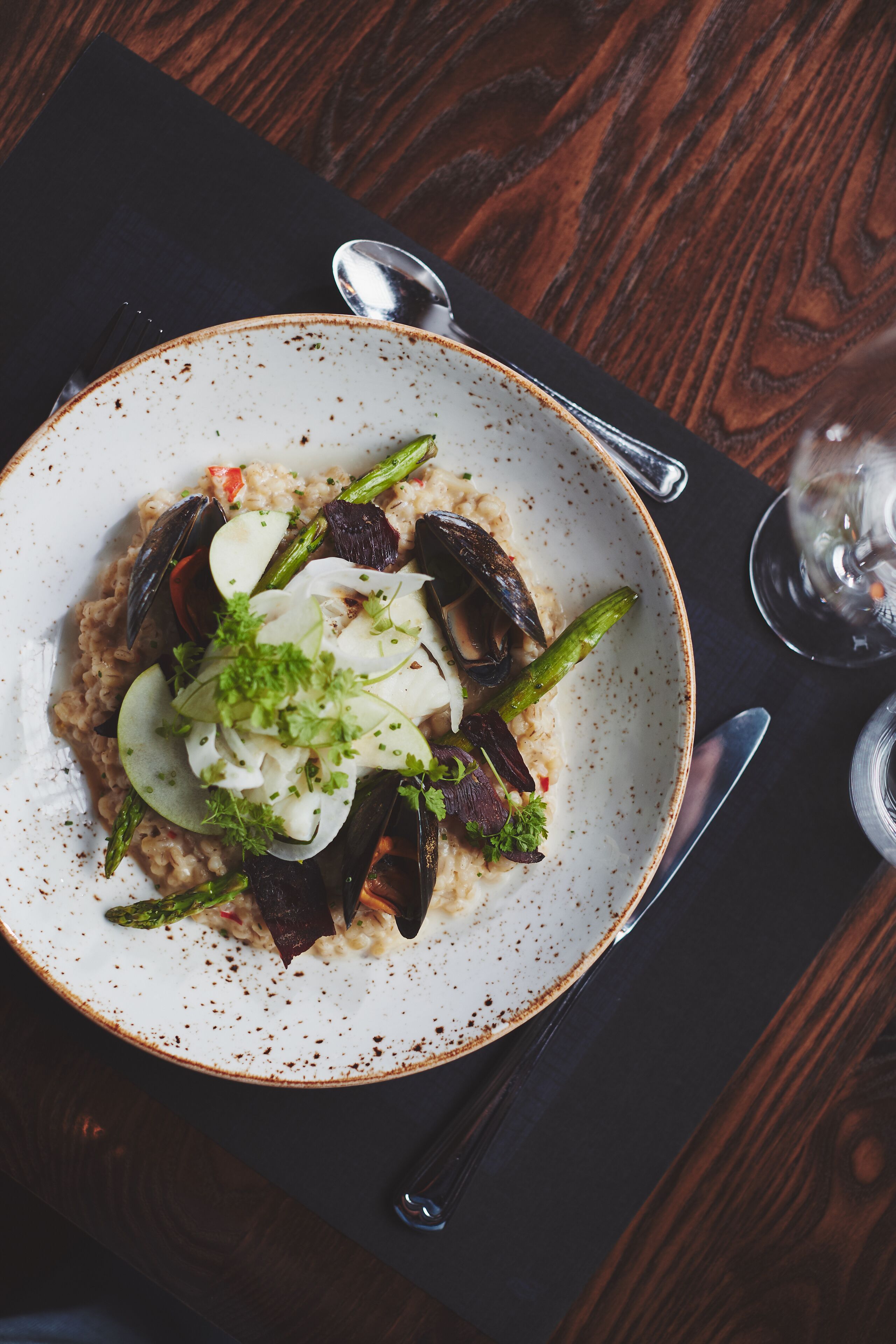 A bowl of food with mussels, asparagus, and a garnish on a plate, placed on a dark wooden table with a spoon and a glass of water nearby.