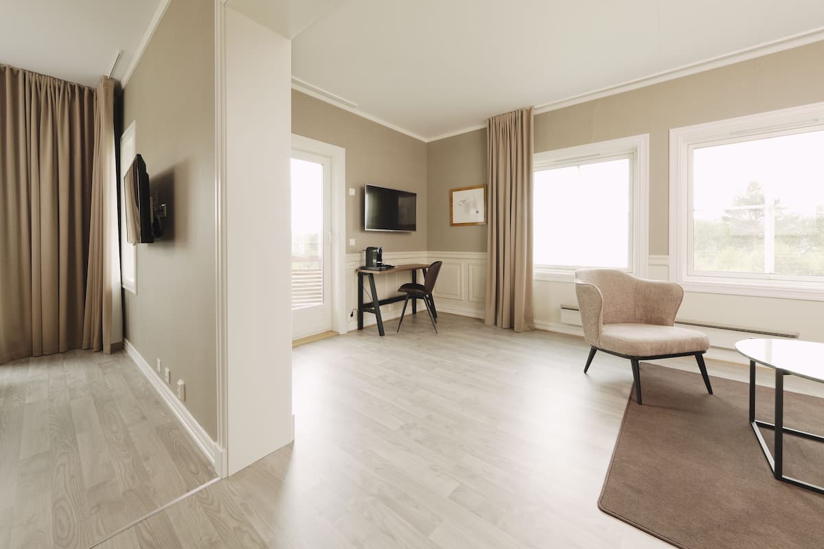 Ein modernes Hotelzimmer mit Holzboden, einem an der Wand montierten Flachbildfernseher, einer Essecke mit Tisch und Stühlen sowie großen Fenstern mit Vorhängen.