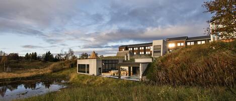 Een modern huis met een grote glazen gevel, een klein buitengebieds keukentje en een vijver op de voorgrond.