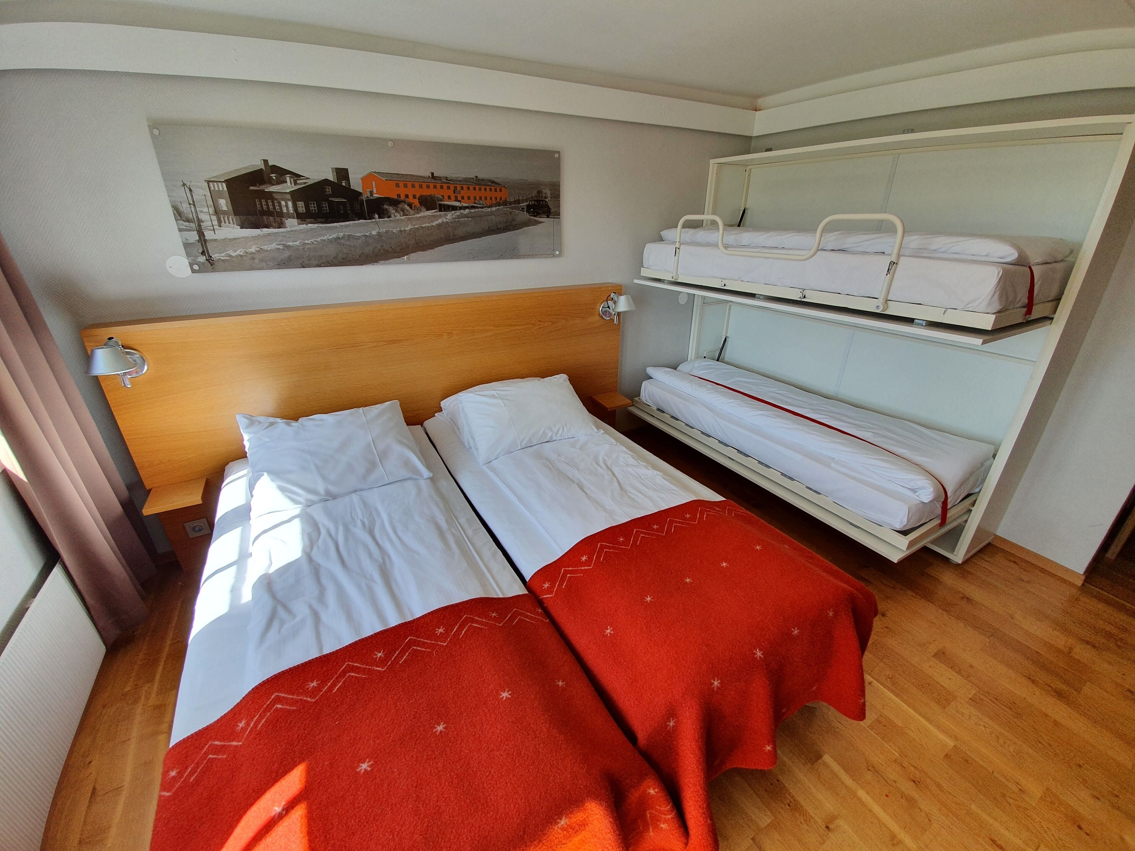 Habitación de hotel con cabecera de madera, dos camas con ropa de cama blanca, una manta roja, un cuadro de un edificio y una litera con ropa de cama blanca.