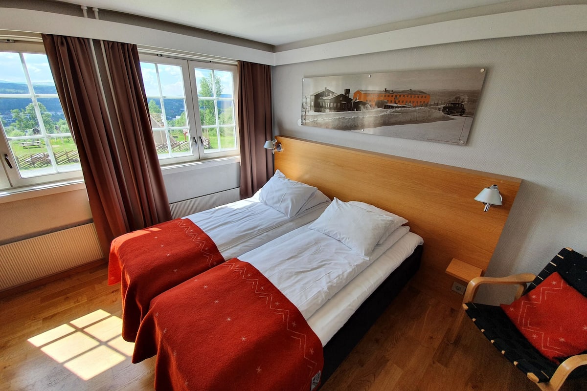 A hotel room with a large bed, two bedside tables, a wooden headboard, a window with curtains, and a painting on the wall.