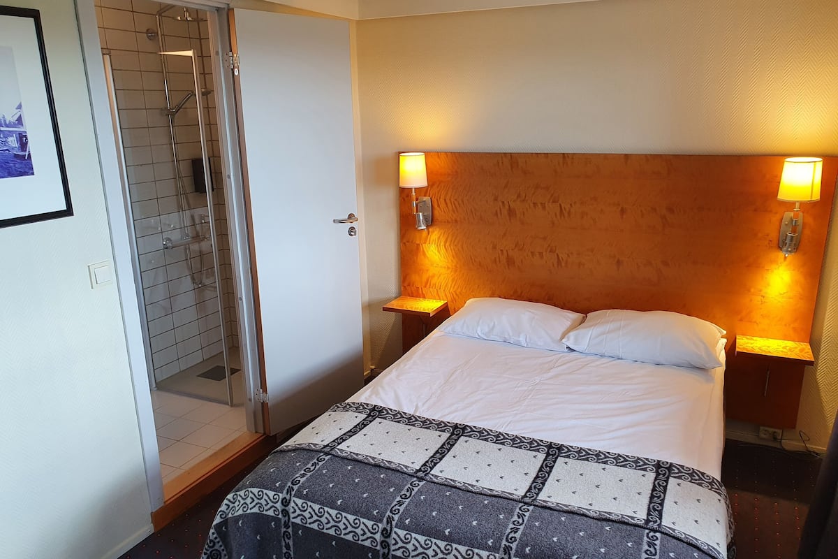 A hotel room with a bed, two bedside tables, a wooden headboard, a shower area, and two wall-mounted lamps.
