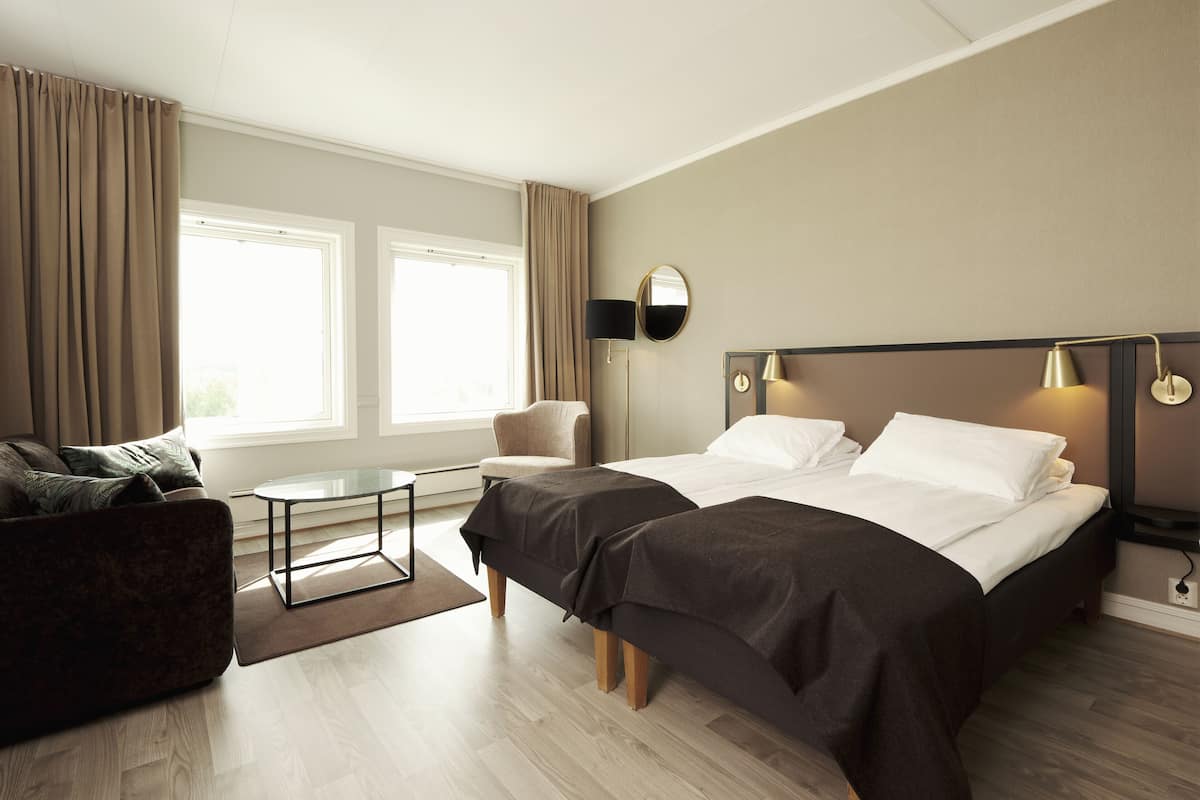A hotel room with a bed, a sofa, a small table, and a chair. There are two windows with curtains, a lamp on the wall, and a mirror.