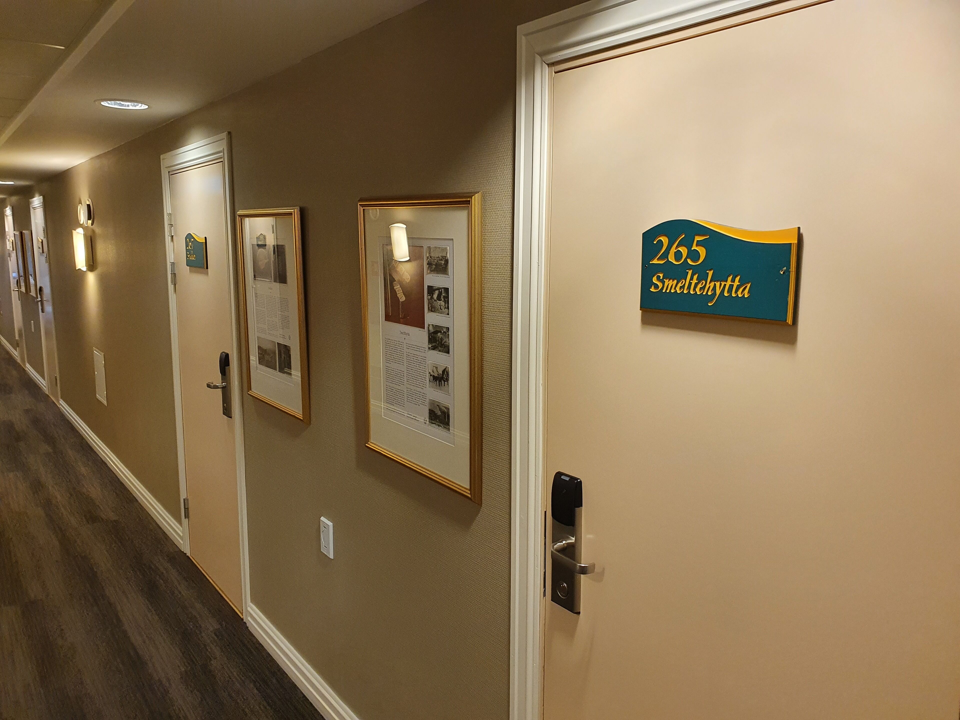 Corredor de hotel com quadros informativos nas paredes, uma porta com placa de número e um interfone.
