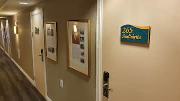 Corredor de hotel com quadros informativos nas paredes, uma porta com placa de número e um interfone.