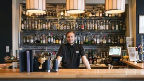 A bar area with a bartender, shelves of liquor bottles, and a cash register. - Roros Hotell (Roros)