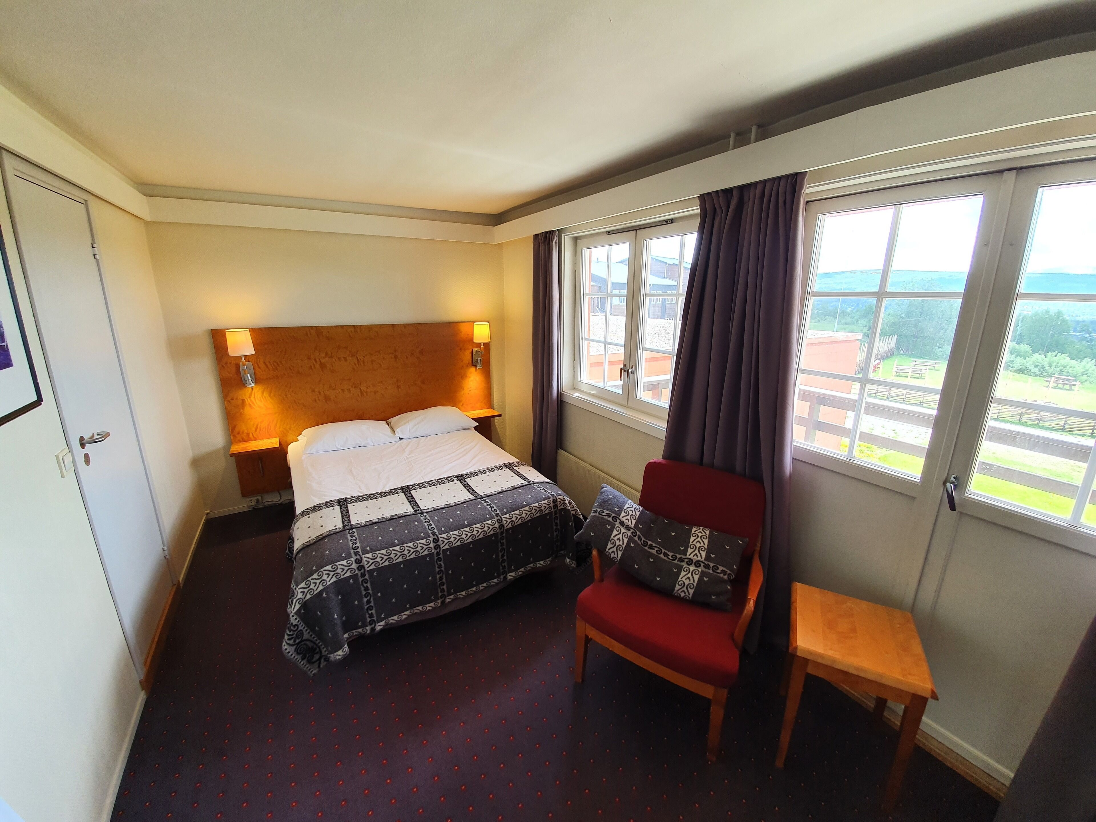 Une chambre d’hôtel avec un lit, une chaise, une petite table, une fenêtre avec des rideaux et une lampe fixée au mur.