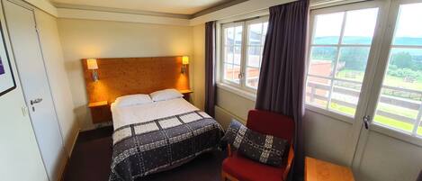 Une chambre d’hôtel avec un lit, une chaise, une petite table, une fenêtre avec des rideaux et une lampe fixée au mur.