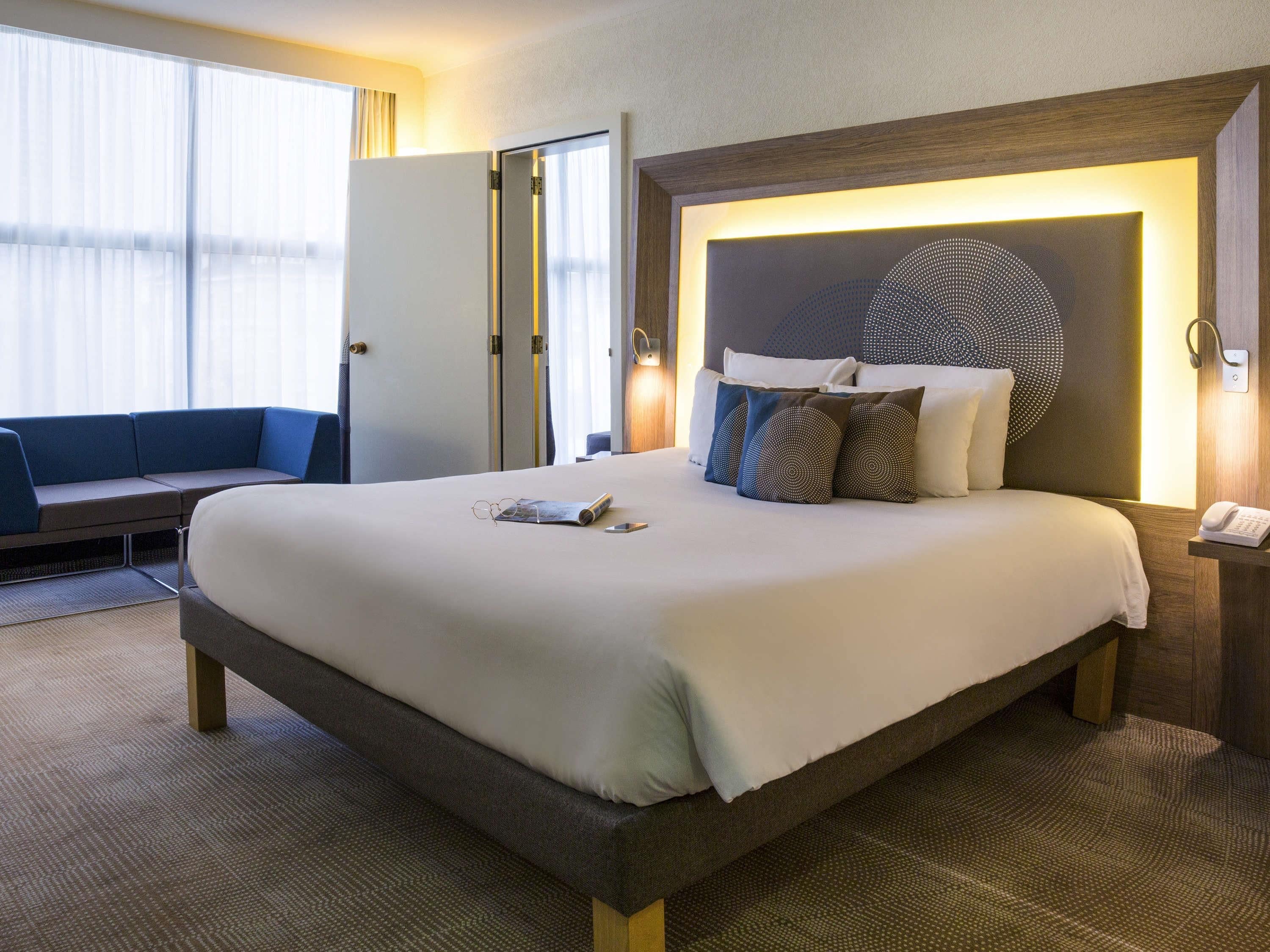 Ein Hotelzimmer mit einem großen Bett, einem Nachttisch mit Telefon, einer Wandleuchte und einer Sitzecke mit Sofa und Vorhängen.